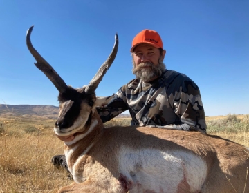 A bearded hunter wearing a camouflage jacket and orange hat sits beside a pronghorn antelope in Wyoming's prairie, showing a successful hunting experience with SNS Outfitter & Guides.